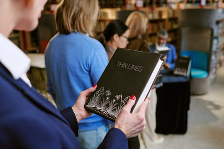 Caucasian middle aged woman holding book titled Thin Lines while standing in line with diverse group of adults at bookstore presentation event, author seated at table in backgroundの写真素材