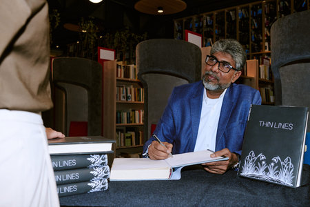 Middle aged man with gray hair and beard sitting at table signing book during presentation event,...