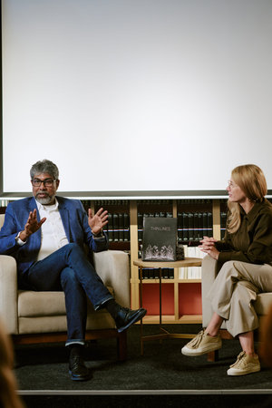 Middle aged Caucasian woman and middle aged South Asian male author sitting in chairs discussing...
