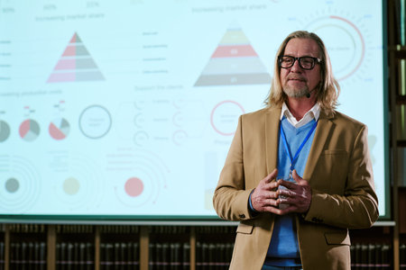 Middle aged man presenting business data in front of large screen with charts and graphs, standing and gesturing during professional presentation in library settingの写真素材