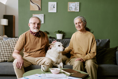 Caucasian senior man and senior woman sitting on sofa with large dog between them, man gently petting dog, both looking at camera, cozy living room background with wall artの写真素材
