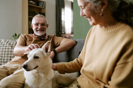 Senior man smiling while petting dog on couch, senior woman sitting beside him also petting dog, both appearing relaxed and enjoying companionship indoorsの写真素材
