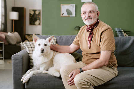 Portrait of senior Caucasian man sitting on sofa with hand resting on dog, both looking at camera, relaxed posture, indoor setting with home decor in backgroundの写真素材