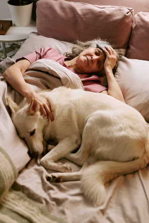 Senior Caucasian woman with headache lying in bed while petting large dog resting beside her,...