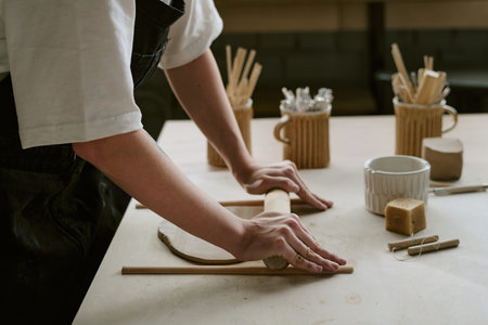 Caucasian young adult woman shaping clay with wooden rolling pin on worktable, hands pressing down...