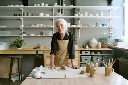Portrait of senior Caucasian woman standing at pottery studio table smiling at camera, hands resting...