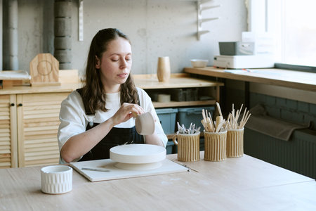 Caucasian young adult woman shaping ceramic cup at pottery wheel, sitting at wooden table with...
