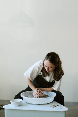 Young adult woman shaping clay on pottery wheel, concentrating on hands while creating ceramic...
