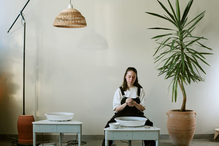 Caucasian young adult woman sitting between tables shaping clay in minimalist indoor setting,...