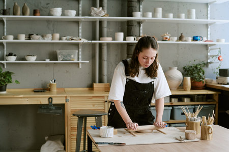 Young adult Caucasian woman shaping clay with rolling pin on wooden table in pottery studio,...