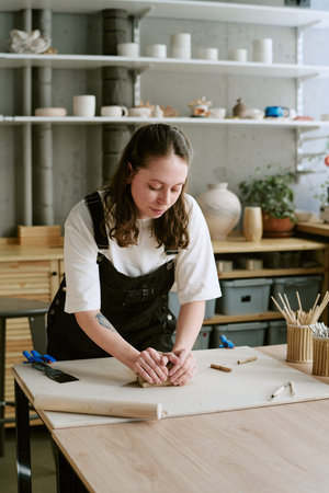 Young adult Caucasian woman shaping clay with hands on worktable in pottery studio, concentrating on...