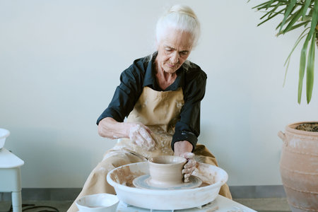 Senior Caucasian woman shaping clay on pottery wheel, concentrating on crafting ceramic bowl with...