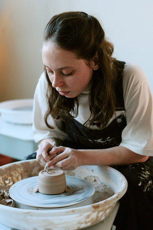 Young adult Caucasian woman shaping clay on pottery wheel, concentrating on sculpting ceramic piece...