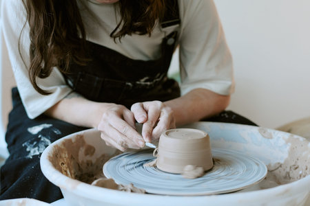 Caucasian young adult woman shaping clay on pottery wheel, hands carefully molding spinning vessel,...