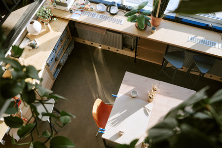 Top view showing empty workspace with wooden tables, chairs, potted plants and art supplies arranged...
