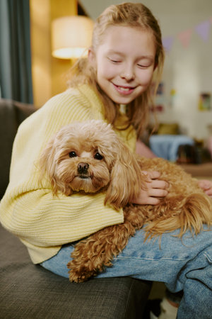 Vertical of girl child smiling while sitting on sofa holding small fluffy dog on lap, looking down...
