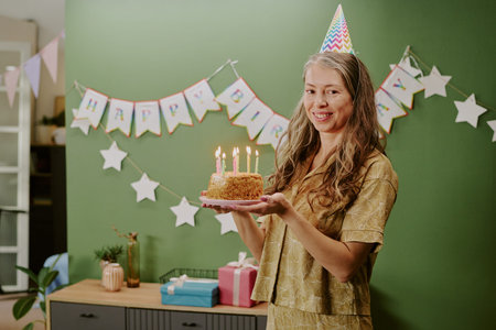 Portrait of middle aged Caucasian woman smiling and holding birthday cake with lit candles while...