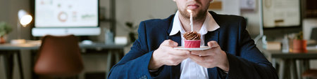 Header of Caucasian young adult man holding plate with cupcake and lit candle, sitting at desk in...