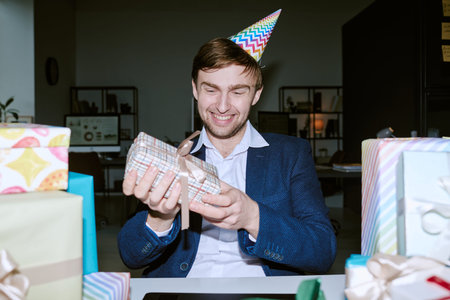 Caucasian young adult man wearing party hat smiling while holding wrapped gift box, surrounded by multiple presents on table, celebrating birthday in modern office environmentの写真素材