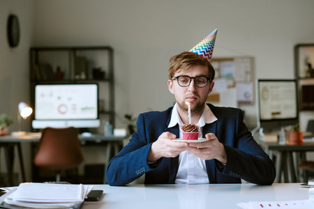 Portrait of Caucasian young adult man wearing party hat sitting at desk holding cupcake with lit...