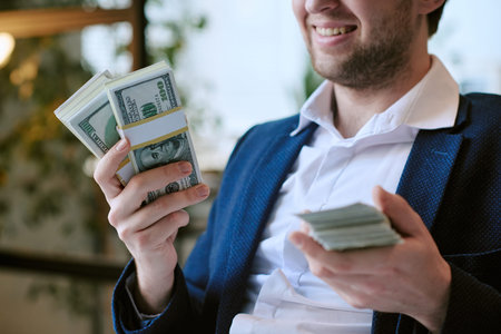 Caucasian young adult man smiling while holding large stacks of US dollar bills in both hands,...