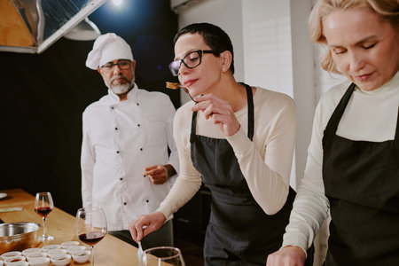 Middle aged woman tasting food while standing beside Caucasian woman preparing ingredients and...