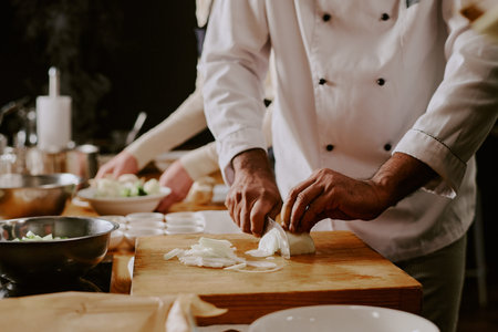 Middle aged man wearing chef uniform slicing onions on wooden cutting board during cooking class,...