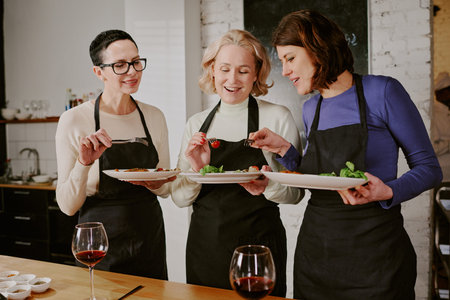 Three middle aged Caucasian women wearing aprons participating in cooking class, holding plates with...