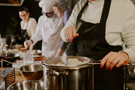 Middle aged Caucasian woman straining cooked rice over pot in foreground, multiethnic group of adults and chef preparing food together in background during cooking classの写真素材