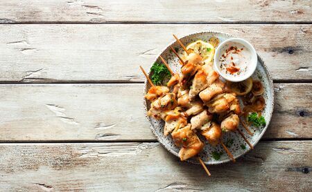 Chicken kebabs on skewers and savory cream sauce on grunge plate on natural wooden table. Flat lay....