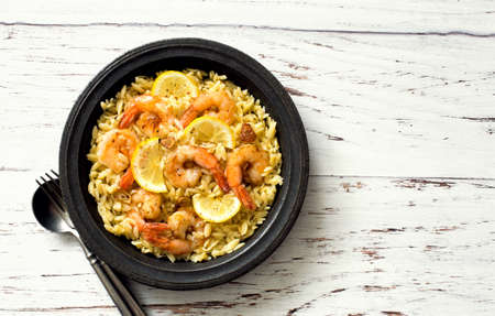 Orzo pasta with shrimps and lemon served on black rustic plate on white wooden  background. Flat lay. Copy spaceの写真素材