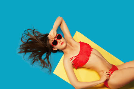 Beach Vacation abstract concept. Colorful studio portrait of young Fashion Woman in red swimsuit and sunglasses. Isolated on Blue background like sea waterの写真素材
