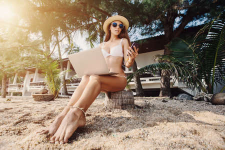 Woman sitting on sea shore working with laptop computer and mobile phone on tropical island beach under palm treeの写真素材
