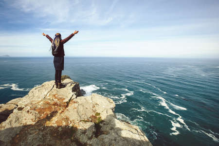 Young female with dreadlocks hair style staying outdoor on Atlantic ocean cliff, keep hands up and enjoy freedomの写真素材