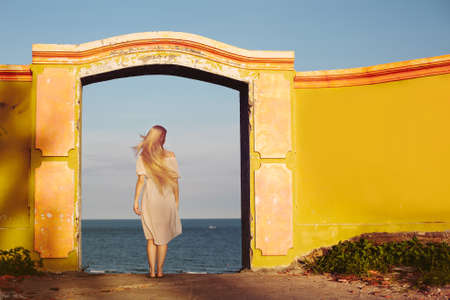 Young Female in white dress staying near arch architecture door with sea view. Beautiful summer day, freedom of choiceの写真素材