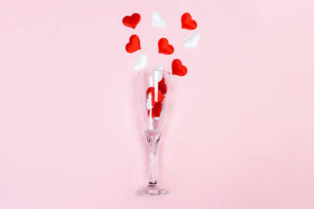 One glass of champagne with a splash of red and white heart shaped confetti on a pink background Valentines day