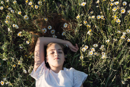 Beauty portrait of a young girl with long hair enjoying nature outdoors, on a field with daisies. Chamomile flowers are woven into long hair. Beautiful young girl face close up.の写真素材