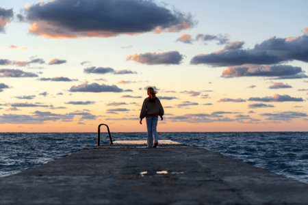 Young woman standing on a pier near the water in the sunset rays against the backdrop of beautiful bright cloudsの写真素材