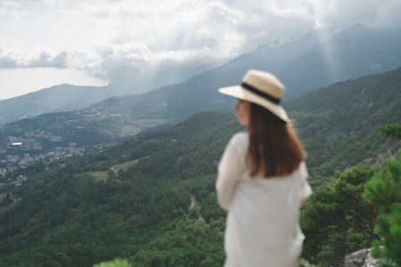 Young beautiful woman with long hair stands with her back raising her hands up, a beautiful view of the mountains and the forest. Travel and healthy lifestyleの写真素材