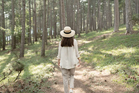 Back view of a young woman walking in a summer forest in white clothes and a straw hat in the sun....