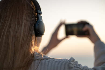 Beautiful young girl walks at sunset near sea makes a self-portrait on smartphone and listens to music on headphonesの写真素材