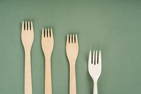 Wooden forks, spoons and one plastic fork on a green background. Eco-friendly disposable tableware....