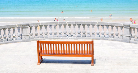 empty modern wooden bench in front of antique stone fence facing the summer beach and blue sea in san sebastian or donostia, basque country, spain, selective focusの写真素材