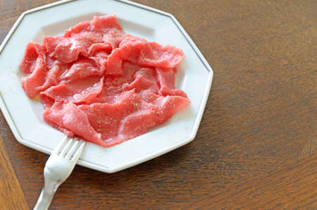 Beef carpaccio with olive oil and fresh ground white pepper on white porcelain plate with silver lining and with stainless steel fork, brown oak wooden backgroundの写真素材