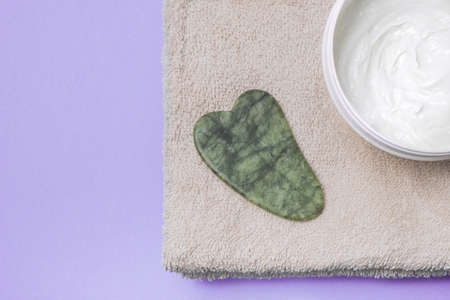 Face care and massage. Face gua sha stone with cream and on a towel on purple backgroundの写真素材