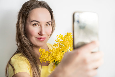 Beautiful young woman with mimosa flowers taking a selfie on white backgroundの写真素材