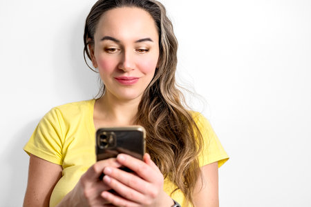 Smiling young woman in blank yellow t-shirt using smartphone on light background. Technology and people conceptの写真素材