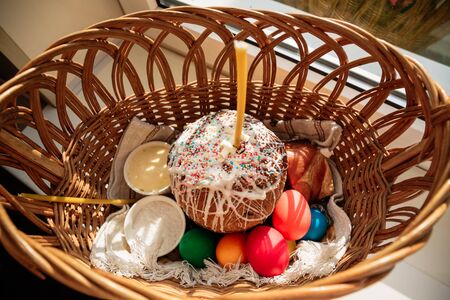 Easter straw basket decorated with a festive napkin with Easter cake and bright decorated eggs and a lit church candle. A festive treat and a peaceful mood.の写真素材