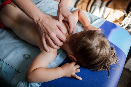 Doctor osteopath massage therapist is doing massage to a little boy. Hands massage the back and cervical-collar zone. Close-up view without face. Mom gives her baby a massage.の写真素材