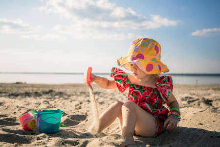 A little cute girl of three years old in a red dress and a panama hat sits on the beach and plays in the sand with plastic toys: a bucket, molds and a shovel. Summertime trip. Family holiday. Close-upの写真素材